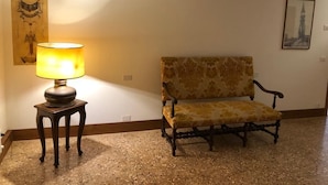 Una habitación con un sofá estampado, una lámpara de pie, una pared con un cuadro enmarcado y un piso con textura.