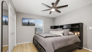 A bedroom with a bed, a ceiling fan, a window with blinds, a wall-mounted TV, and a door.