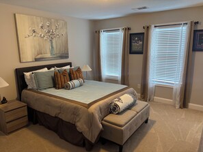 A bedroom with a large bed, a nightstand, a bench, a painting, and two windows with blinds.