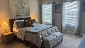 A bedroom with a large bed, bedside lamps, a bench, and two windows with blinds.