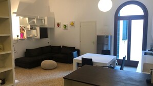 A modern living space with a dark-colored sofa, a white dining table, and a kitchen area with a refrigerator. - Rio Carmini (Venice)