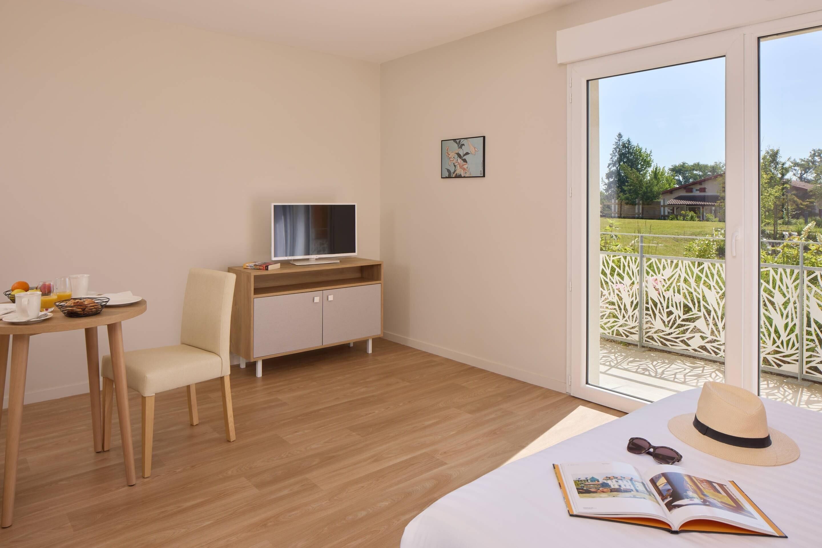 Une chambre d’hôtel comprenant un lit, un bureau avec un ordinateur, une chaise, une table à manger avec des articles pour le petit-déjeuner, et une grande fenêtre donnant sur un espace verdoyant et un immeuble.