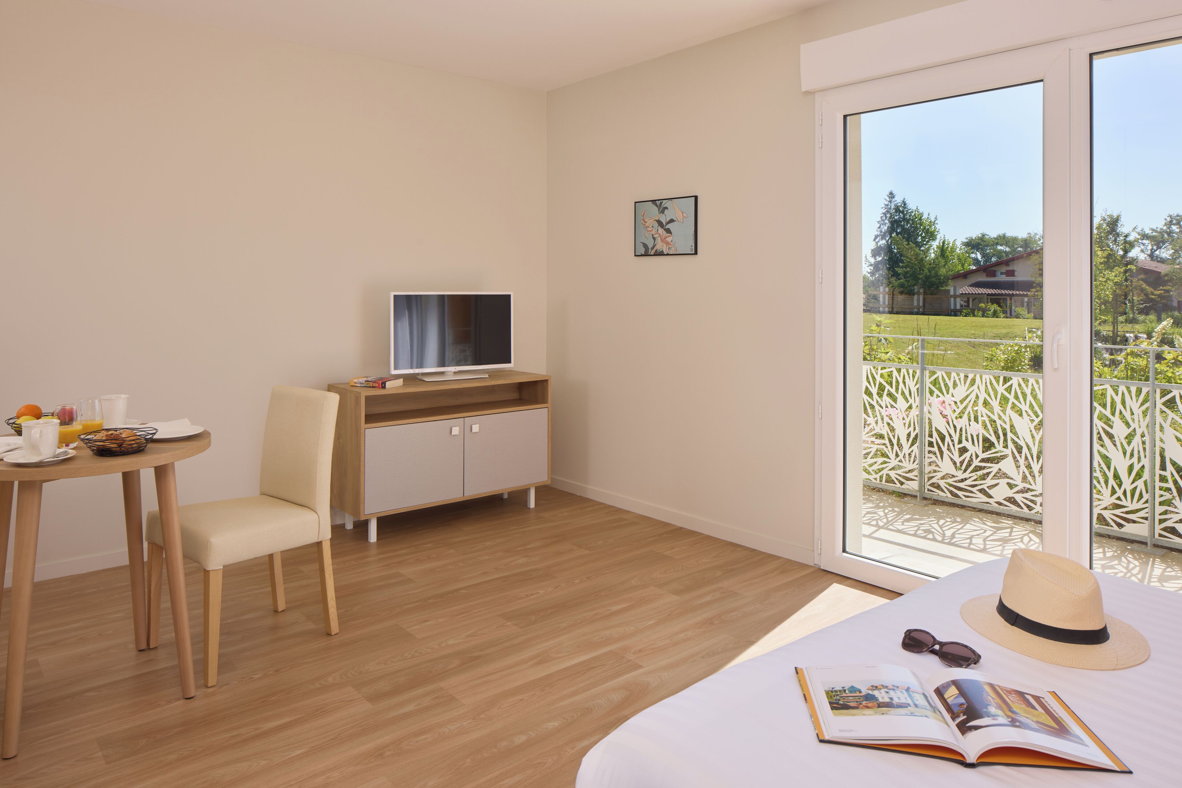 A hotel room with a bed, a desk with a computer, a chair, a dining table with breakfast items, and a window with a view of greenery.