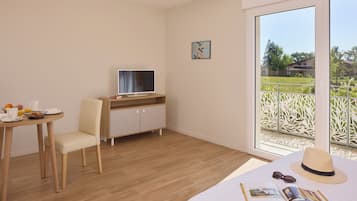 Ein Hotelzimmer mit Bett, Schreibtisch mit Computer, Stuhl, Esstisch mit Frühstücksangeboten und einem Fenster mit Blick auf Grünflächen.