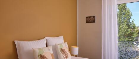 A hotel room with a large bed, two pillows, a nightstand with a lamp, and a view of greenery through a window.