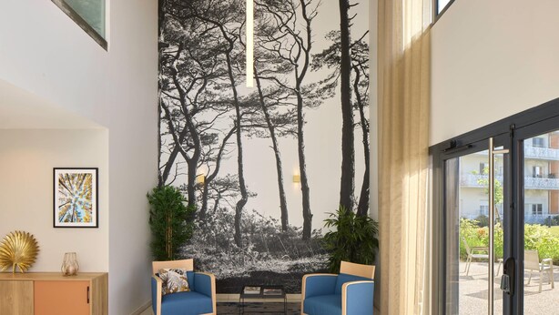 A modern living room with two blue armchairs, a wooden sideboard, a large tree mural on the wall, and a view of an outdoor balcony. - Quality Aparthotel Saint-Paul-lès-Dax (Saint-Paul-lès-Dax)