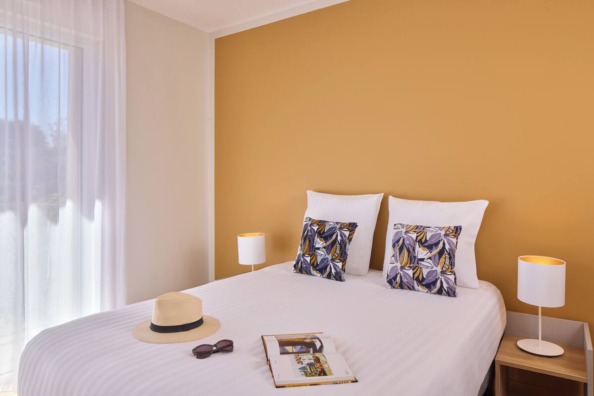 Ein Hotelzimmer mit einem großen Bett, zwei Kissen, einem Hut, einer Sonnenbrille und einer Zeitschrift auf dem Bett.