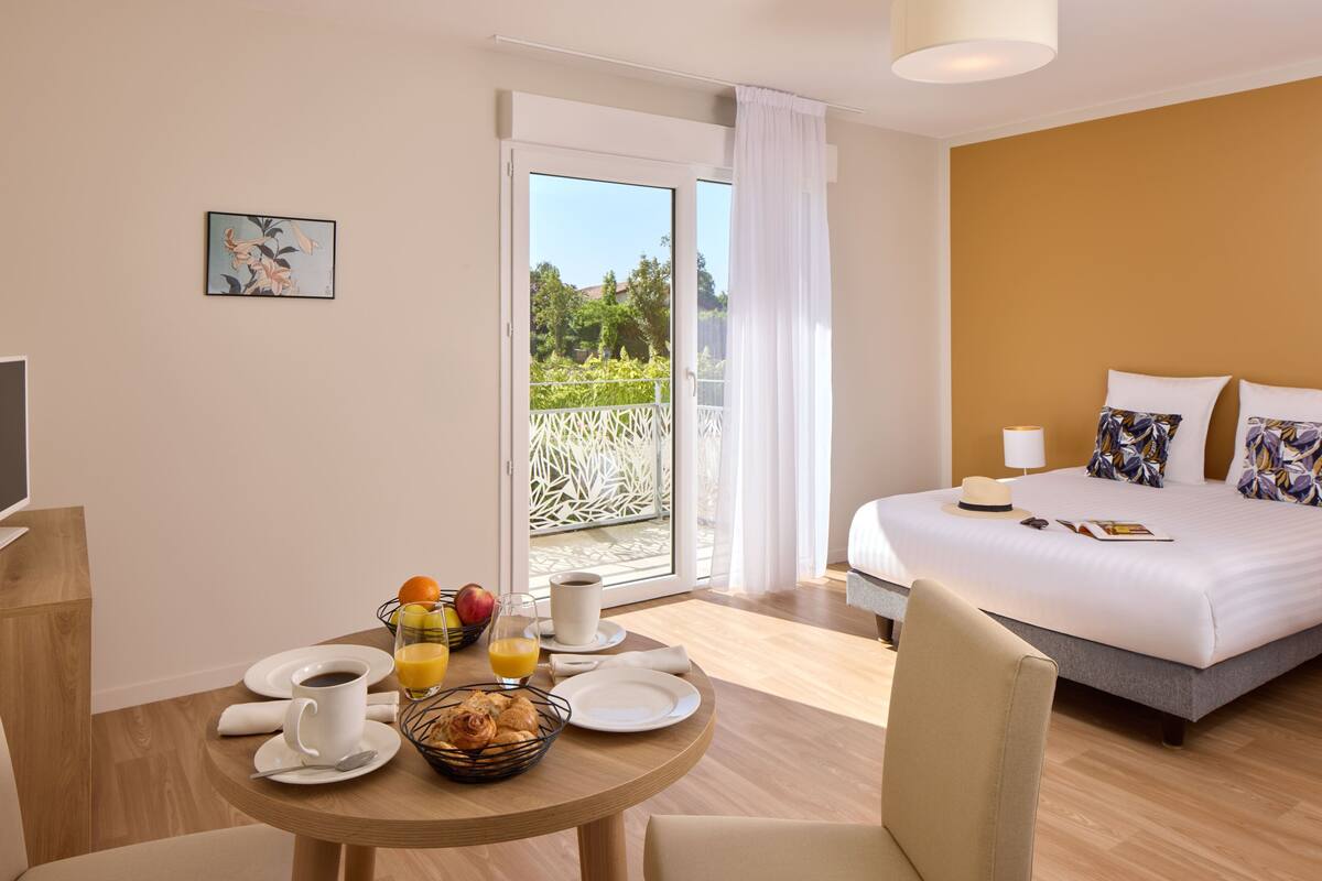 Ein Hotelzimmer mit Bett, Frühstückstisch mit Obst, Kaffee und Blick auf Grün.