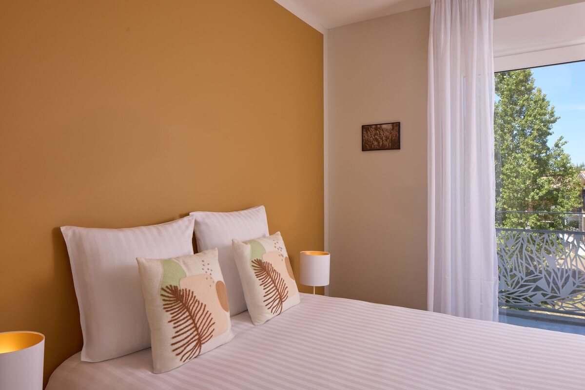 A hotel room with a bed, pillows, a nightstand, a lamp, and a view of the outdoors.