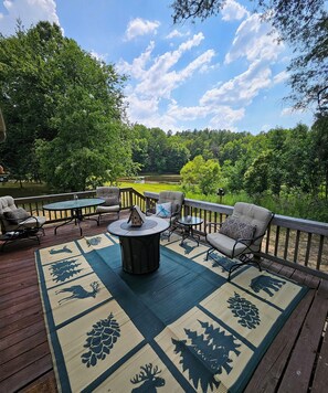 A deck with outdoor furniture, a fire pit, and a view of a lake surrounded by trees.