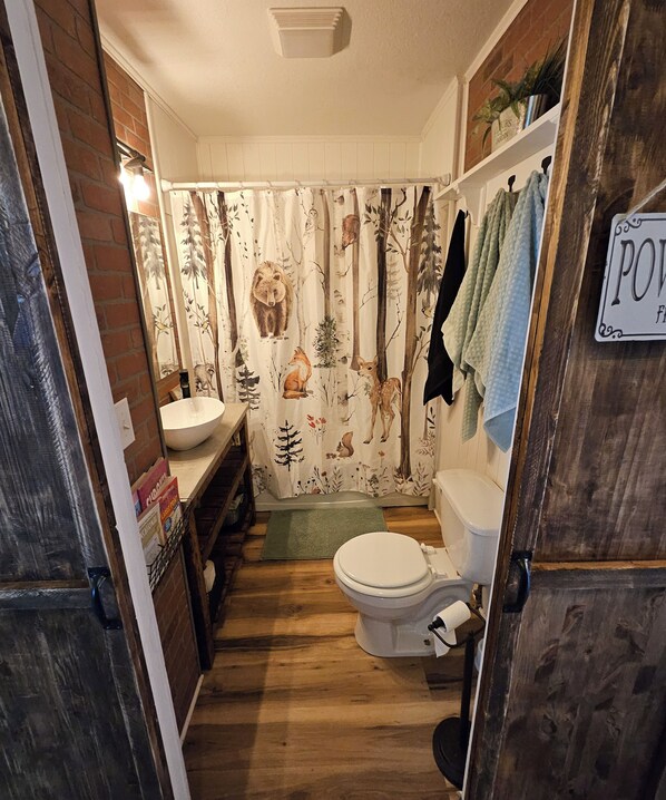 A bathroom with a rustic design, featuring a wooden floor, a white toilet, a sink, and a shower curtain with forest animal prints.