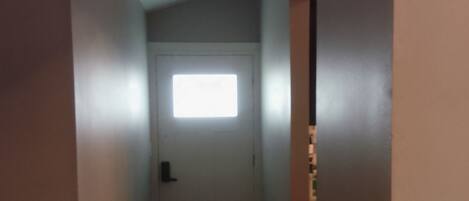 Un pasillo estrecho con una puerta al final, una ventana pequeña y suelo de madera.