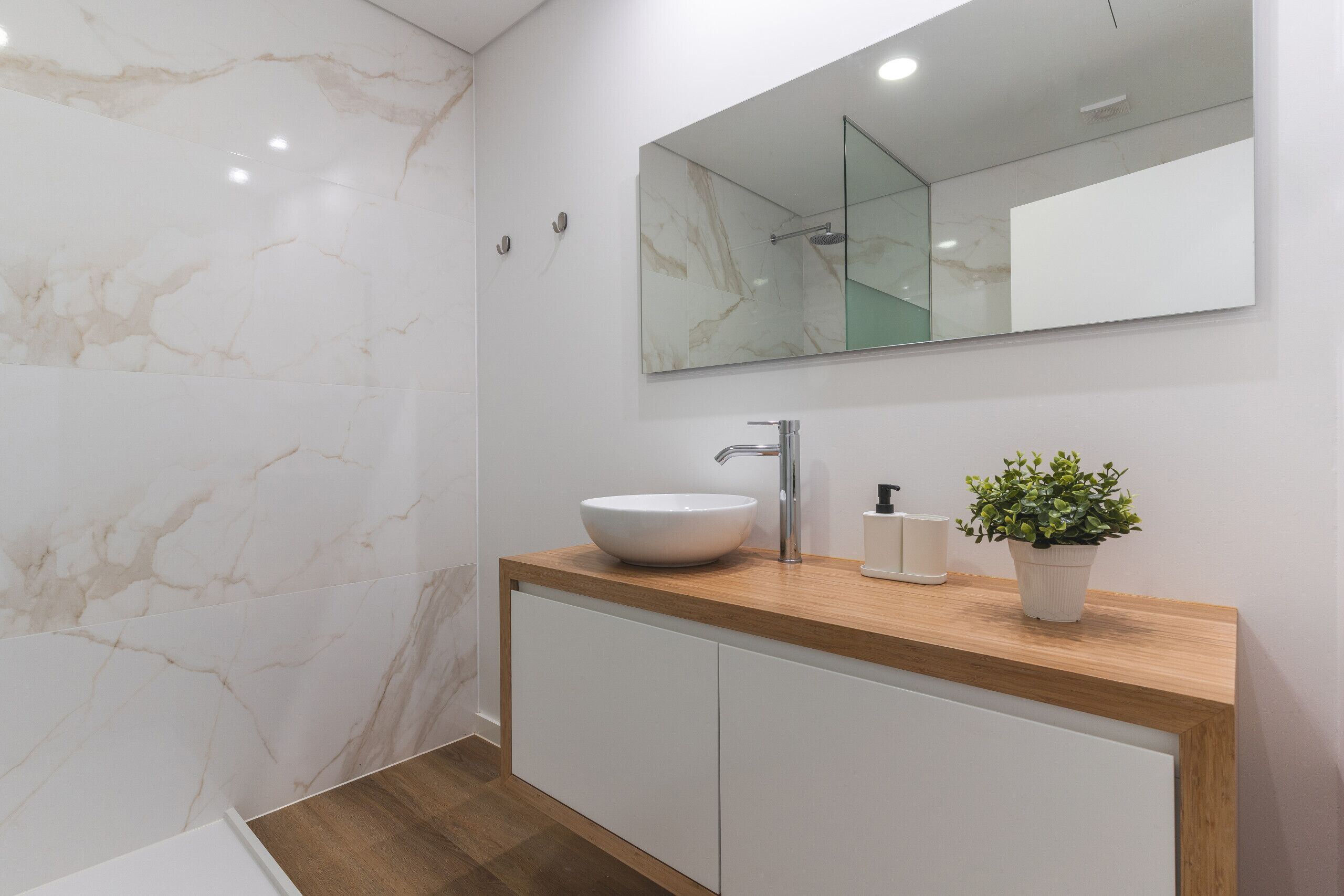A bathroom with a marble wall, a wooden vanity, a white sink, a mirror, and a potted plant.