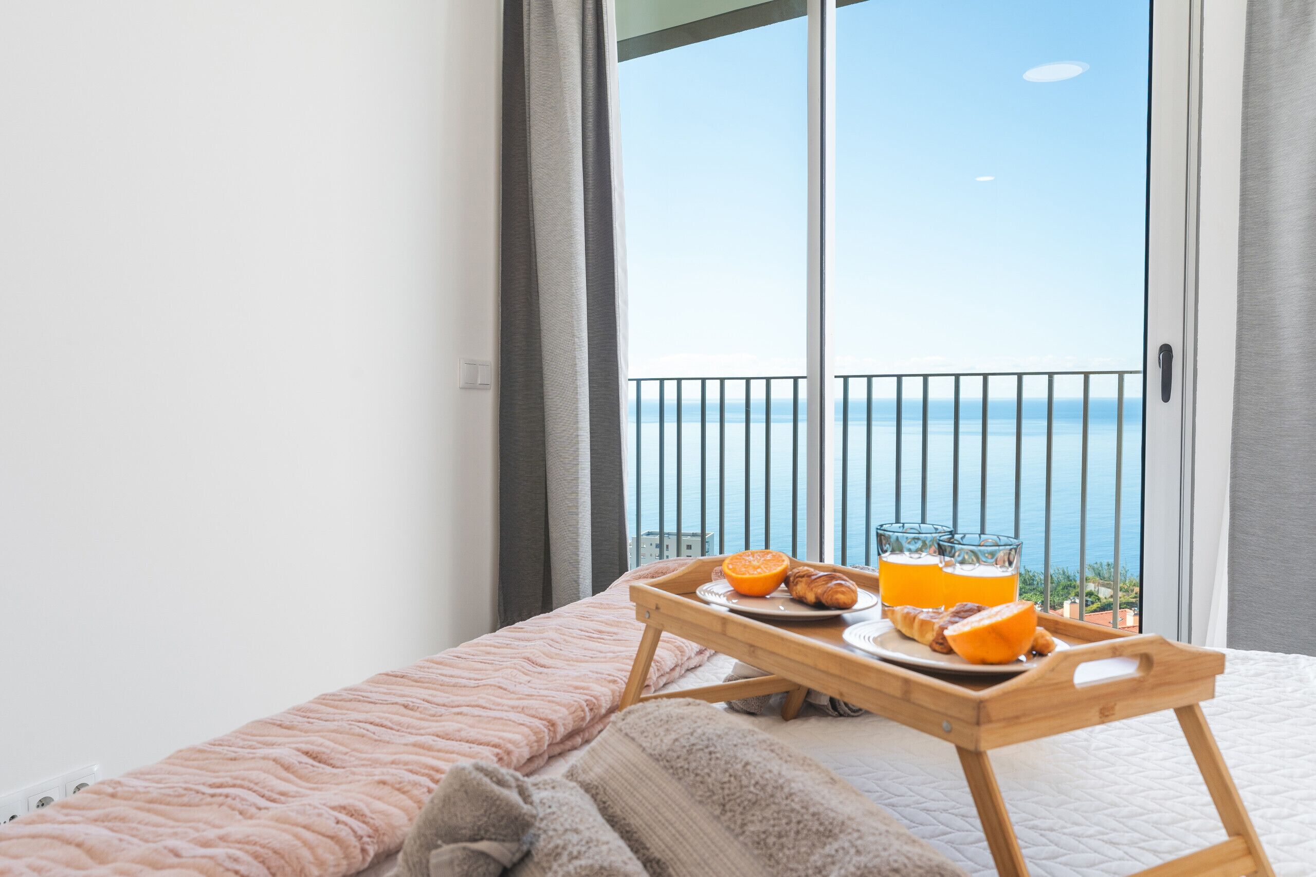 A balcony with a breakfast tray containing croissants, orange slices, and orange juice, overlooking the sea.