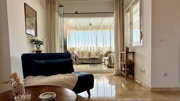 A living room with a sofa, a coffee table, and a dining table. There is a window with curtains, a chandelier, and a view of another room.