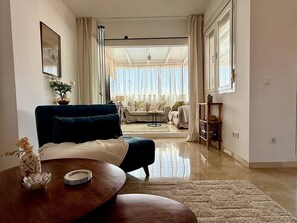 Ein Wohnzimmer mit einer Couch, einem Couchtisch und einem Esstisch. Es gibt ein Fenster mit Vorhängen, eine Kristallleuchter und Blick in einen weiteren Raum.