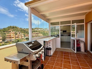 Uma varanda com churrasqueira, uma porta de correr de vidro que dá para uma cozinha e vista para uma área residencial montanhosa.
