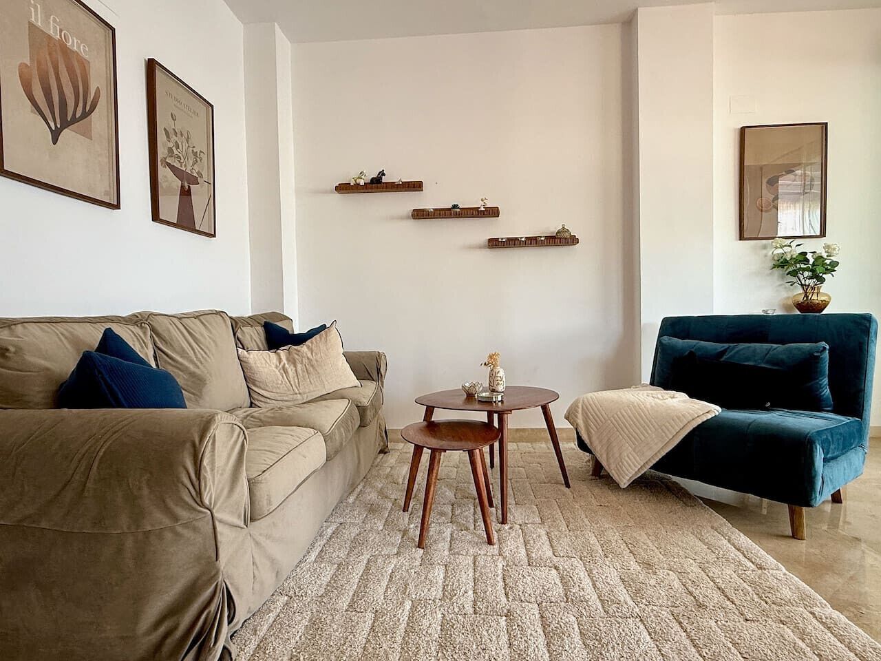 Uma sala de estar com um sofá bege, uma poltrona azul, uma mesa de madeira pequena e um tapete estampado.