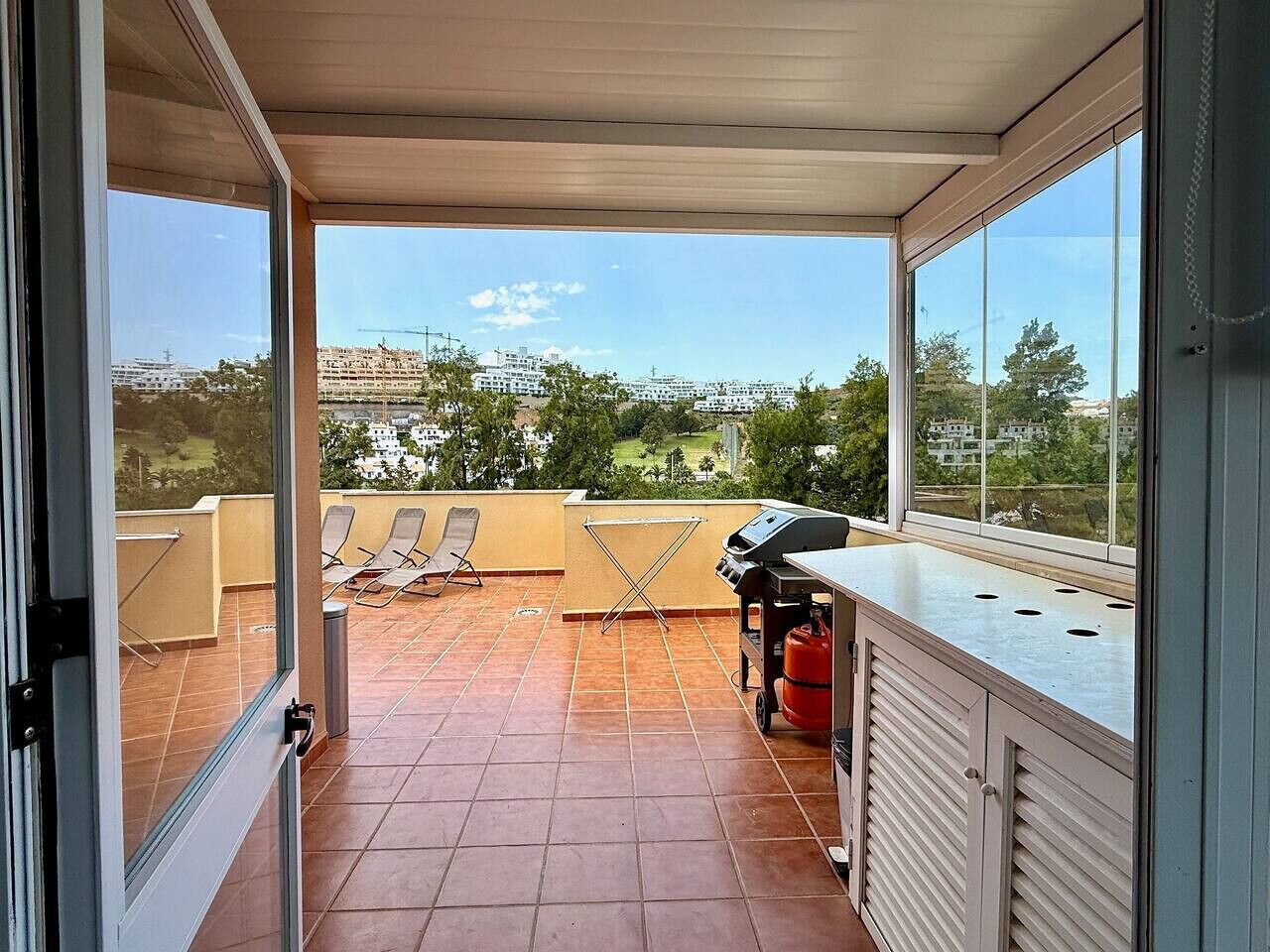 A balcony with a grill, chairs, and a view of buildings and greenery.
