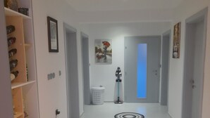 Un pasillo con suelo blanco, una puerta azul al final y un cubo de basura. Se observan patrones circulares en el suelo y una estantería con botellas a la izquierda.