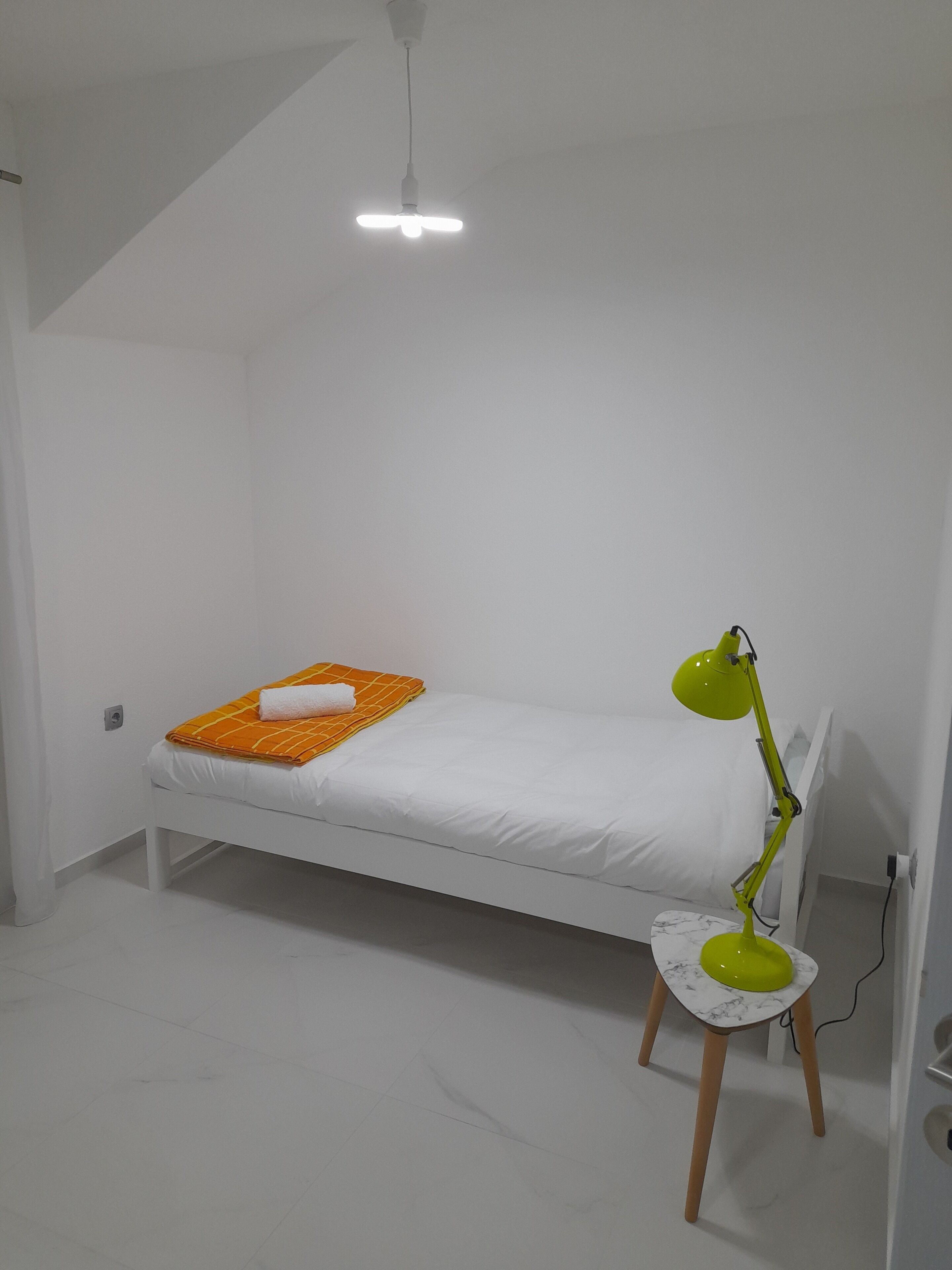 Ein kleines, weißes Zimmer mit einem Einzelbett, Deckenventilator, einer grünen Schreibtischlampe und einem Nachttisch mit Marmormuster.