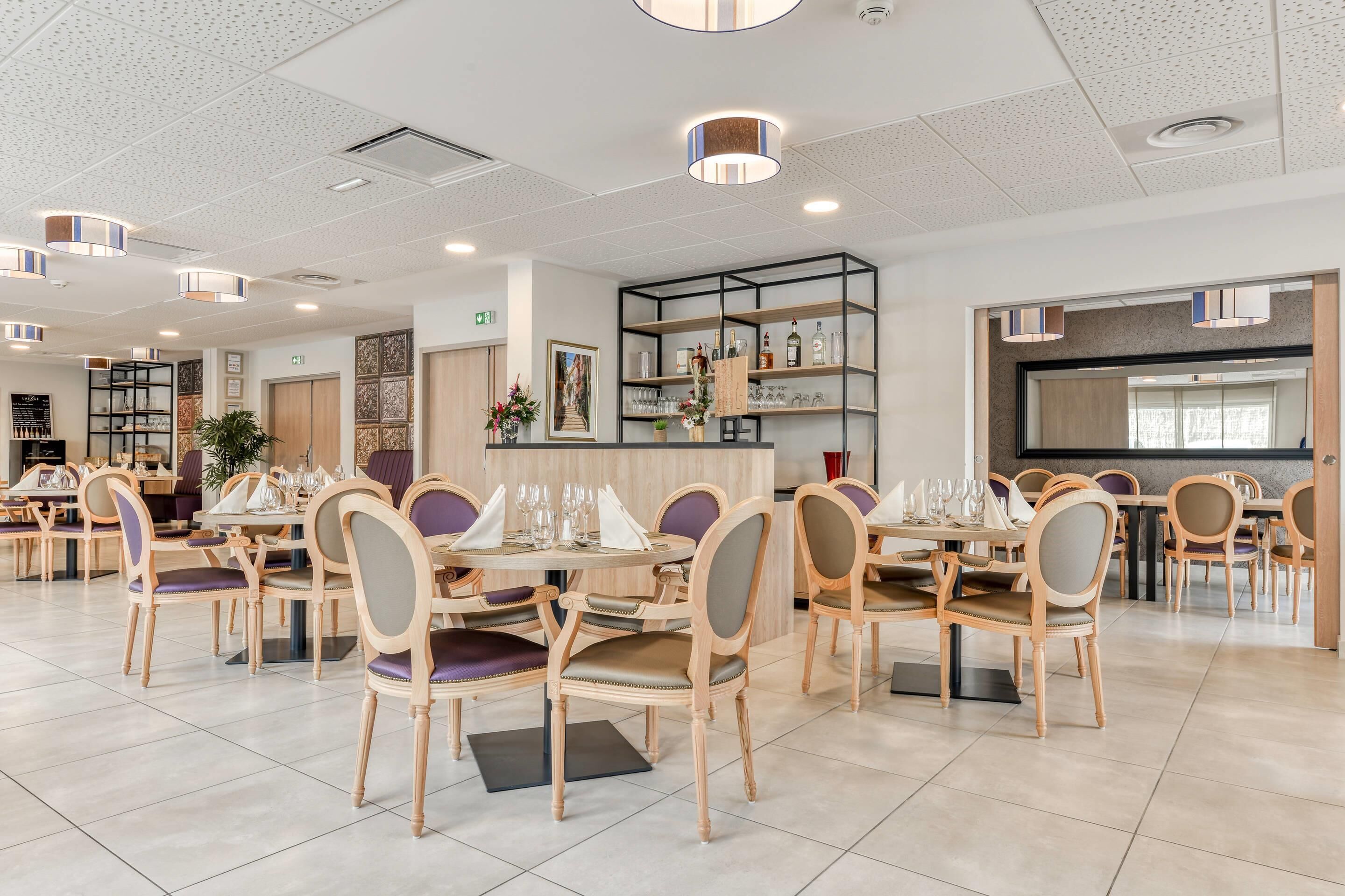 A spacious restaurant with multiple dining tables and chairs, a bar area, and a wall-mounted shelf with bottles.