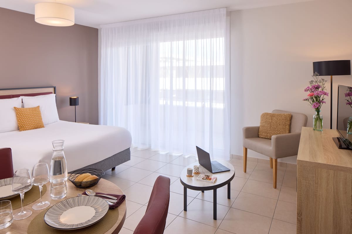 Une chambre d’hôtel comprenant un lit, un bureau, une chaise, une table à manger et une fenêtre avec des rideaux légers.