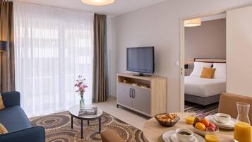 Habitación de hotel con sofá, televisión, mesa de desayuno y cama al fondo.