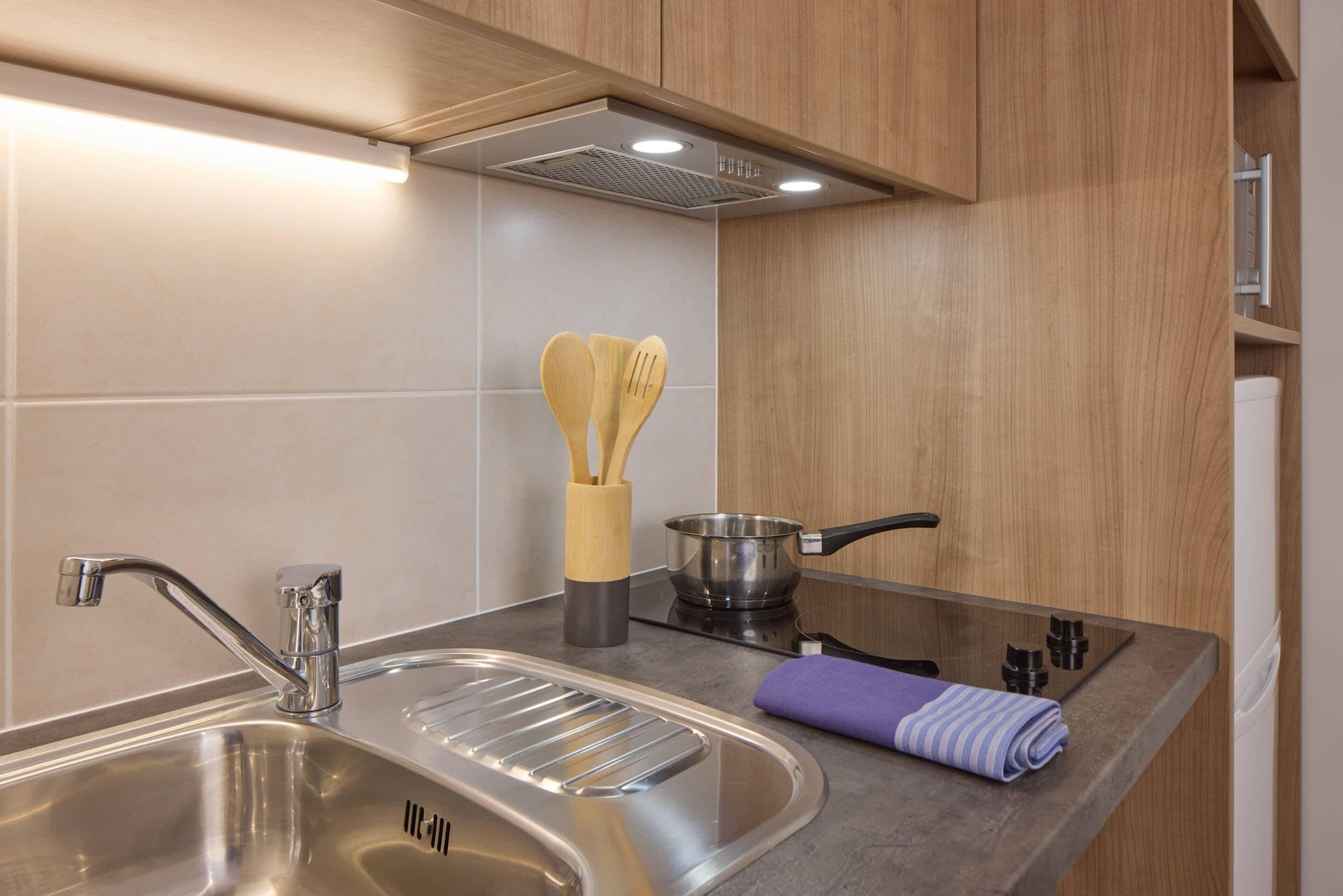 A modern kitchen with a stainless steel sink, a stove, wooden cabinets, and a pot with a spatula.