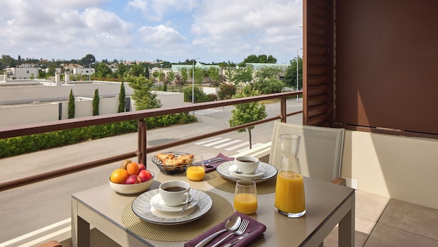 Varanda com mesa de café da manhã, incluindo frutas, café e suco, com vista para a rua e edifícios.