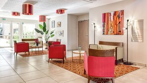 Um hall de hotel moderno com assentos em vermelho e bege, uma planta grande e uma obra de arte colorida na parede.