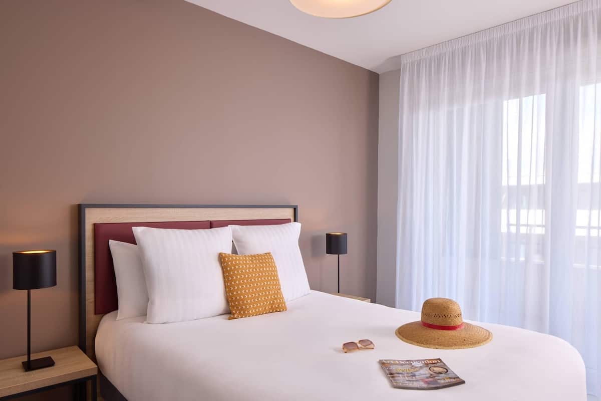 Un lit bien fait, avec des oreillers blancs et jaunes, un magazine et un chapeau de paille posés sur le lit.