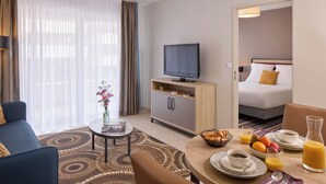 Quarto de hotel com sofá, televisão, mesa de jantar e itens de café da manhã, além de uma cama ao fundo.