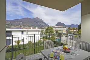 A balcony with a table set for dining, a bowl of fruit, and a view of houses and mountains. - Corte Fasanella appartamento Lillà (Toscolano-Maderno)