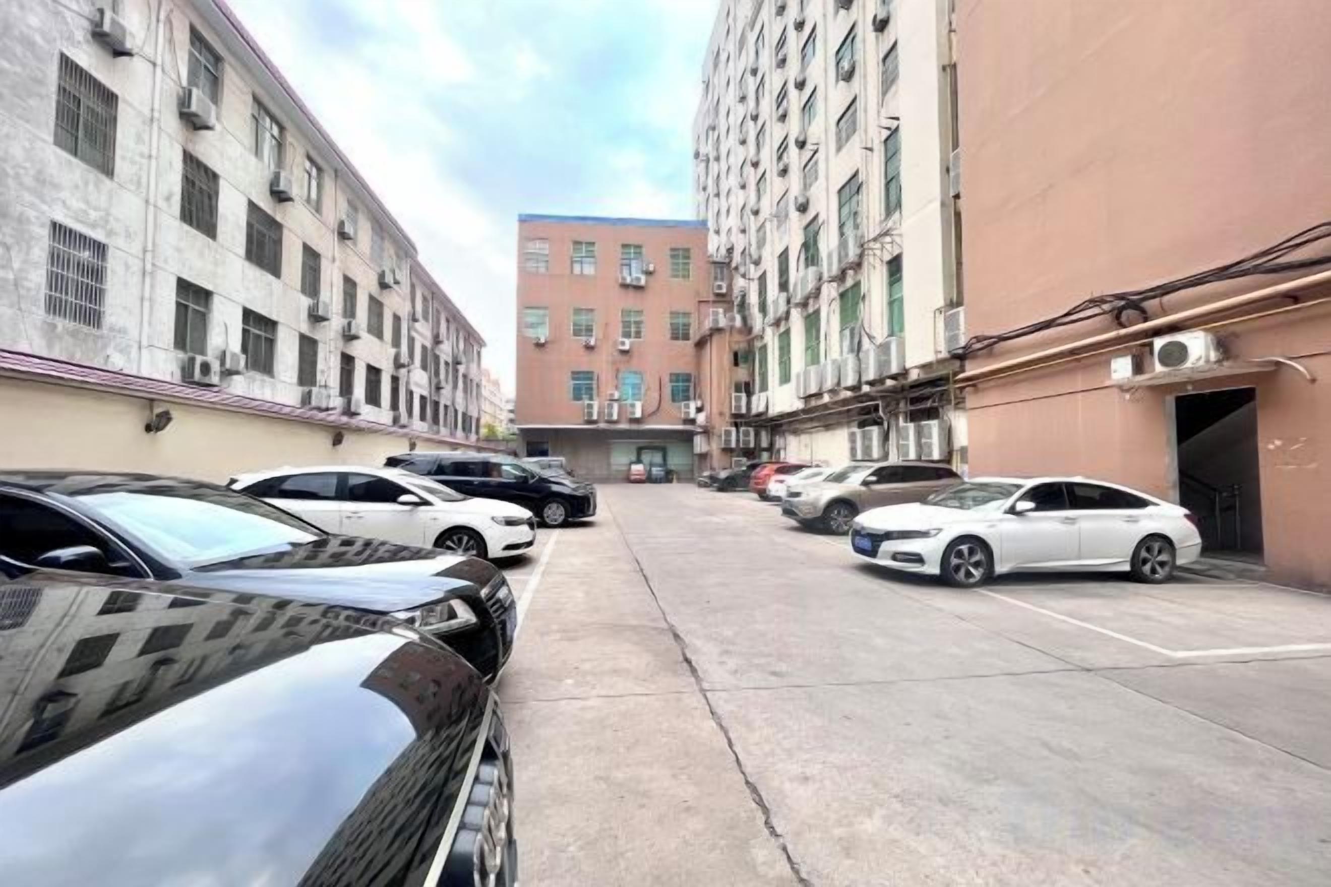 A parking lot with cars parked in rows, surrounded by multi-story buildings with air conditioning units.