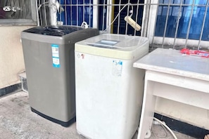 Deux gros appareils électroménagers, probablement un lave-linge et un sèche-linge, sont placés à côté d’une table blanche sur laquelle se trouve un objet rouge.