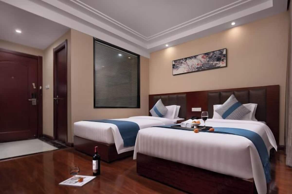 Une chambre d’hôtel avec deux lits, une grande fenêtre avec des stores, un tableau au mur et une bouteille de vin sur la table.