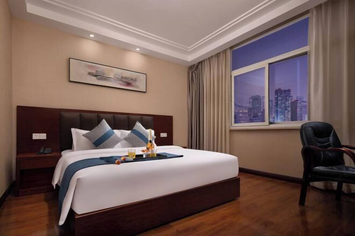 Une chambre d’hôtel avec un grand lit, une table de chevet, une chaise, une vue sur la ville et un tableau encadré au mur.