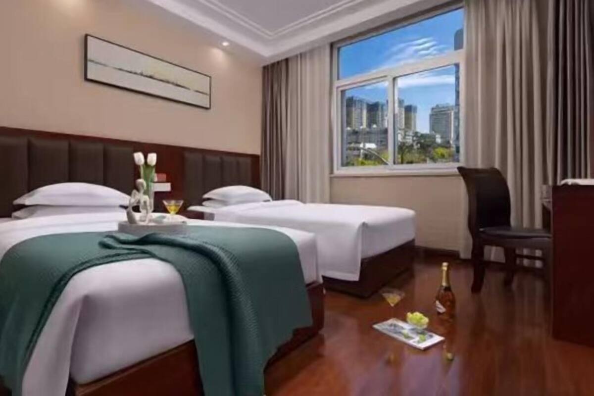 Une chambre d’hôtel avec deux lits, un bureau, une chaise, une fenêtre donnant sur la ville et un tableau encadré au mur.