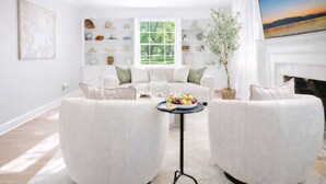 A spacious living room with a white sofa set, a coffee table with fruit, a fireplace, and a large window with a view of greenery.