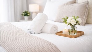 A neatly made bed with rolled white towels, a lit candle, and a vase of white flowers on a tray.