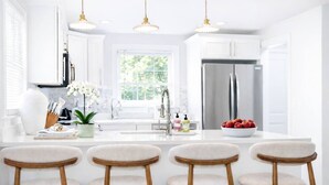 A modern kitchen with white cabinetry, a central island with bar stools, stainless steel appliances, and a large window providing natural light.