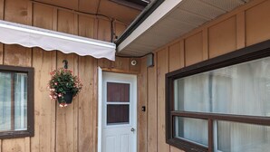 A wooden exterior with a white door, a window with a white awning, and a hanging flower basket.