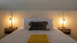 Una cama bien hecha con ropa blanca y una única almohada oscura, flanqueada por dos mesitas de noche con lámparas, en una habitación de diseño sencillo y limpio.