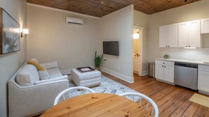 Ein modernes Wohnzimmer mit einer Couch, Hocker und einem Esstisch. Es gibt eine Küchenzeile mit weißen Schränken und einem Geschirrspüler. Ein Flachbildfernseher ist an der Wand befestigt.
