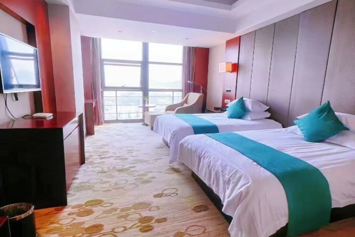 A hotel room with two beds, a TV, a desk, and a large window.