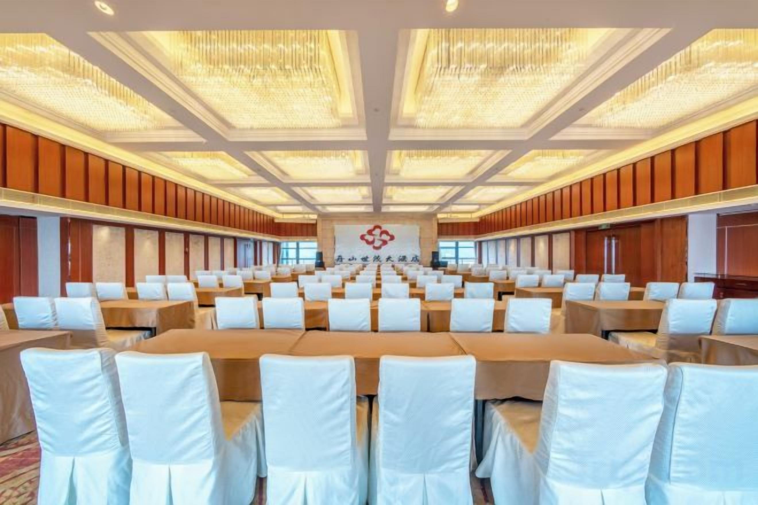 A conference room with rows of tables and chairs, a chandelier, and a logo on the wall.