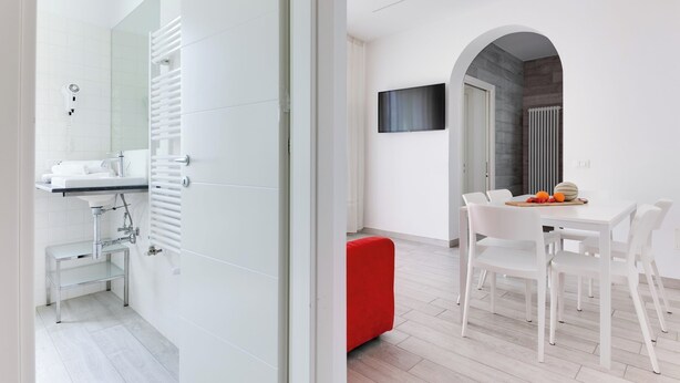A modern bathroom with a white sink, mirror, and towel rack. A red bench is visible in the dining area with a table and chairs. - Four-room apartment Scola 01 (Gabicce Mare)