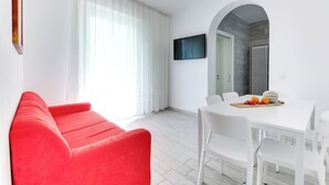 A modern living room with a red sofa, a white dining table with chairs, and a TV mounted on the wall. - Four-room apartment Scola 01 (Gabicce Mare)