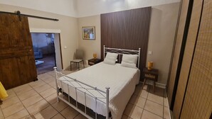 Un dormitorio con estructura de cama de metal blanco, puerta de madera, mesita de noche pequeña con lámpara y una silla.